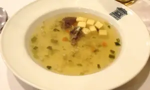 Un bol de potage clair avec des morceaux de viande, des dés de fromage, des légumes verts et des carottes.