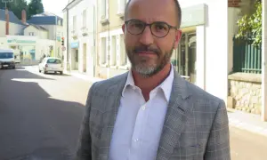 Laurent Raymond, homme politique, porte une veste à carreaux et une chemise blanche, debout dans une rue ensoleillée.