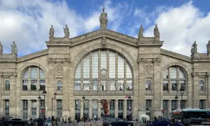 La **gare du Nord** à Paris, majestueuse façade ornée de statues, sous un ciel nuageux. Des voyageurs et des véhicules animent la place.