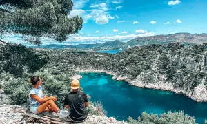 Deux personnes assises sur un tronc d'arbre contemplent une crique aux eaux turquoise, entourée de falaises rocheuses et de pins.