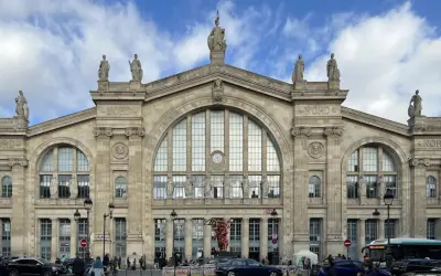 La **gare du Nord** à Paris, majestueuse façade ornée de statues, sous un ciel nuageux. Des voyageurs et des véhicules animent la place.