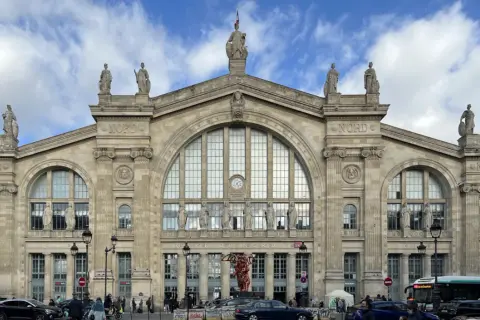 La **gare du Nord** à Paris, majestueuse façade ornée de statues, sous un ciel nuageux. Des voyageurs et des véhicules animent la place.
