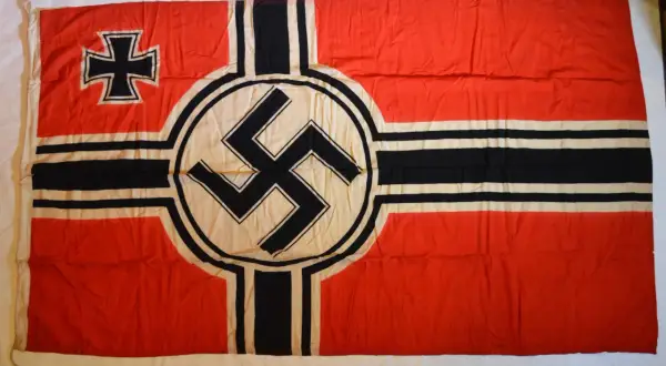Drapeau rouge avec une croix noire et blanche dans le coin supérieur gauche et une croix gammée noire sur un cercle blanc au centre. L'origine de la croix gammée est complexe.