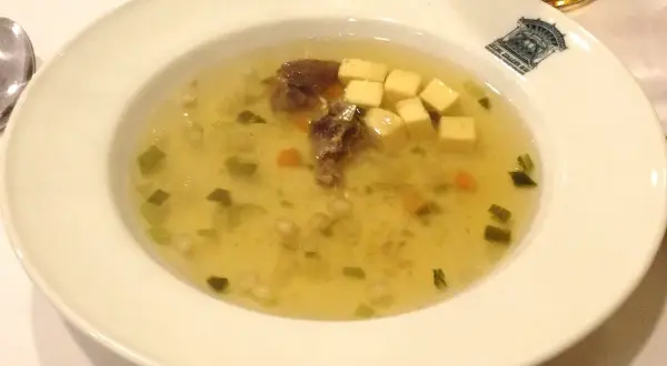 Un bol de potage clair avec des morceaux de viande, des dés de fromage, des légumes verts et des carottes.