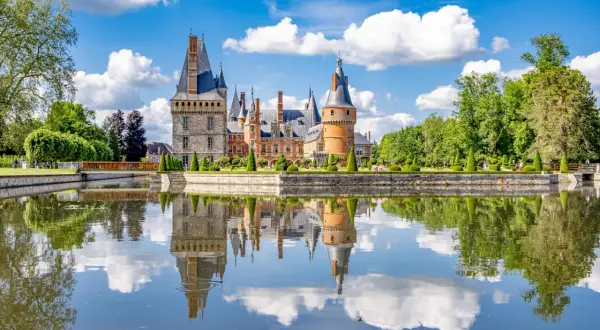 Magnifique château en Eure-et-Loir, reflet parfait dans l'eau calme, entouré de jardins à la française et d'arbres verdoyants sous un ciel bleu parsemé de nuages.