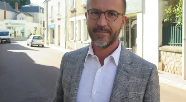 Laurent Raymond, homme politique, porte une veste à carreaux et une chemise blanche, debout dans une rue ensoleillée.