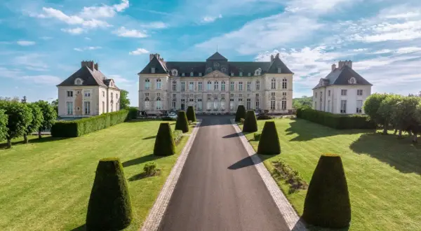Le majestueux château de Brienne, avec ses ailes symétriques et son allée bordée de topiaires, sous un ciel bleu éclatant.
