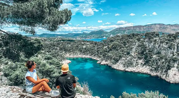 Deux personnes assises sur un tronc d'arbre contemplent une crique aux eaux turquoise, entourée de falaises rocheuses et de pins.