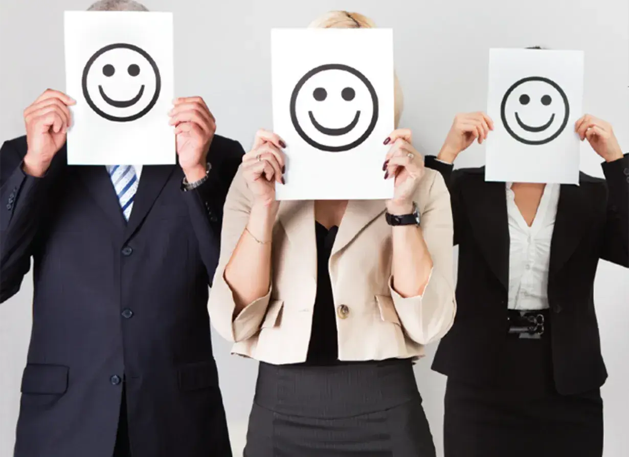 Trois personnes en tenue professionnelle cachent leur visage derrière des feuilles avec des smiley. Le bonheur est une idée neuve en Europe, et ces visages souriants le prouvent.