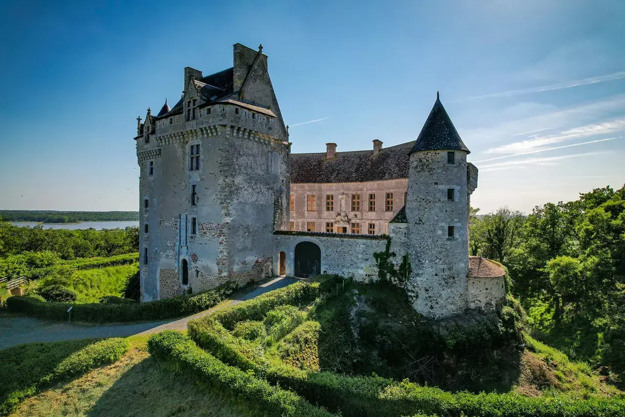 Le majestueux **château du Bouchet** se dresse fièrement sur une colline verdoyante, dominant un lac paisible sous un ciel bleu éclatant.