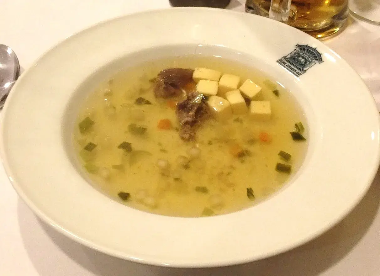 Un bol de potage clair avec des morceaux de viande, des dés de fromage, des légumes verts et des carottes.