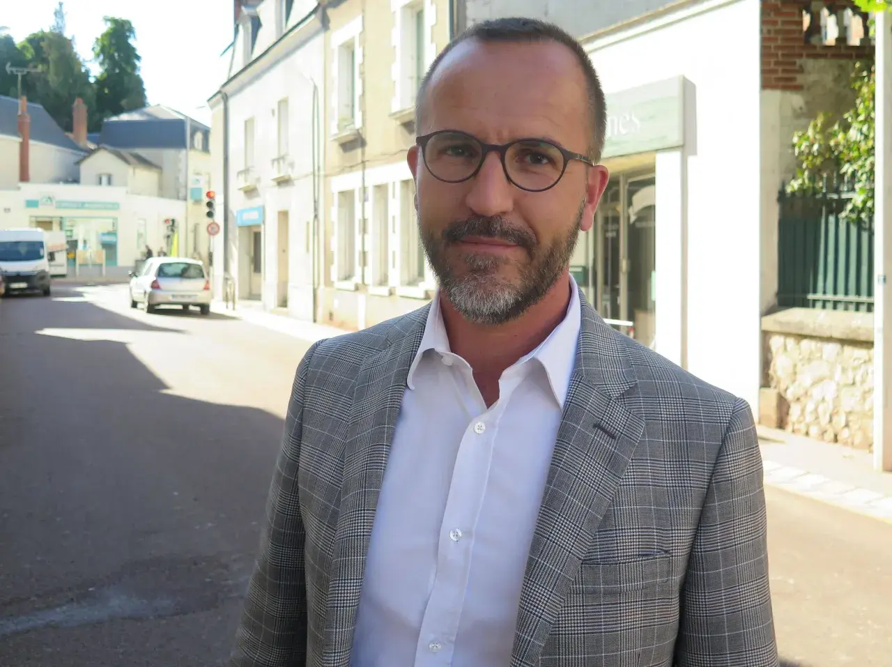Laurent Raymond, homme politique, porte une veste à carreaux et une chemise blanche, debout dans une rue ensoleillée.