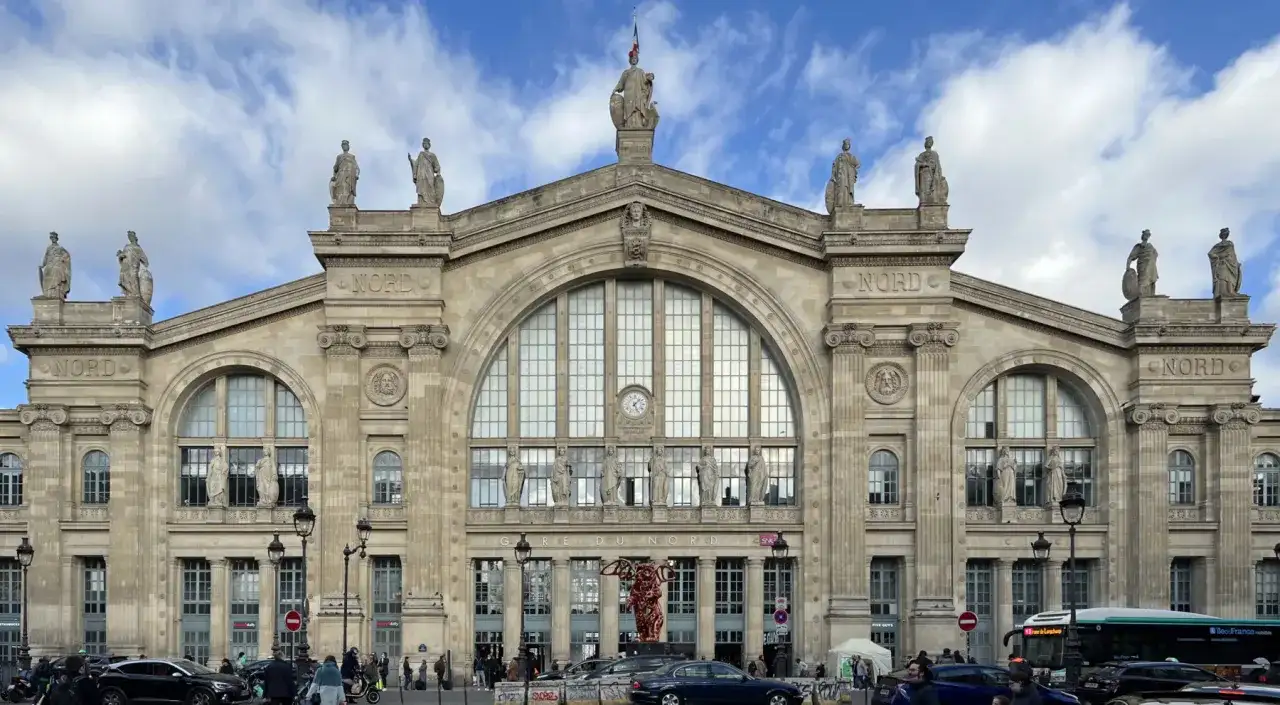 La **gare du Nord** à Paris, majestueuse façade ornée de statues, sous un ciel nuageux. Des voyageurs et des véhicules animent la place.