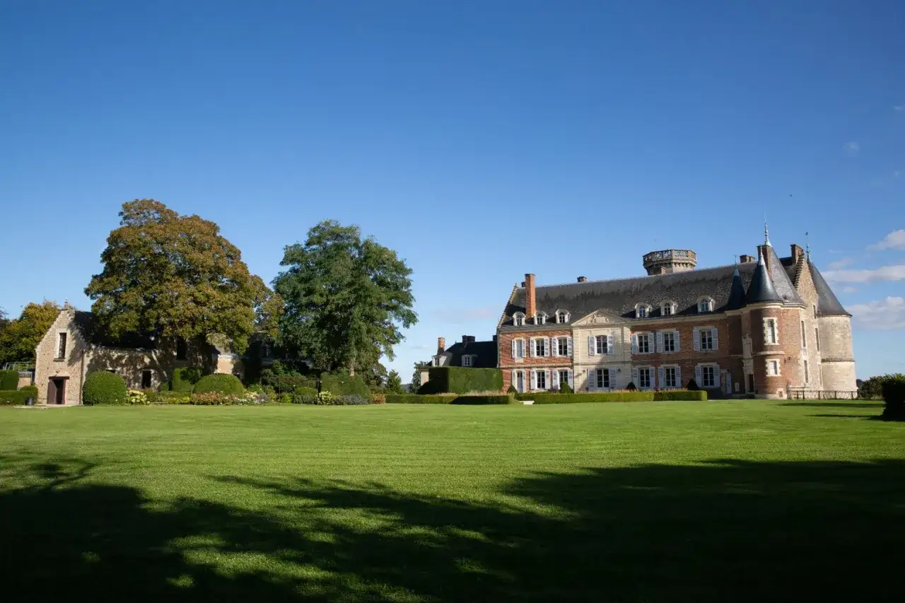 Le château de Montmirail, majestueux édifice de pierre et de brique, se dresse fièrement sous un ciel bleu, entouré d'une pelouse verdoyante et d'arbres aux couleurs d'automne.