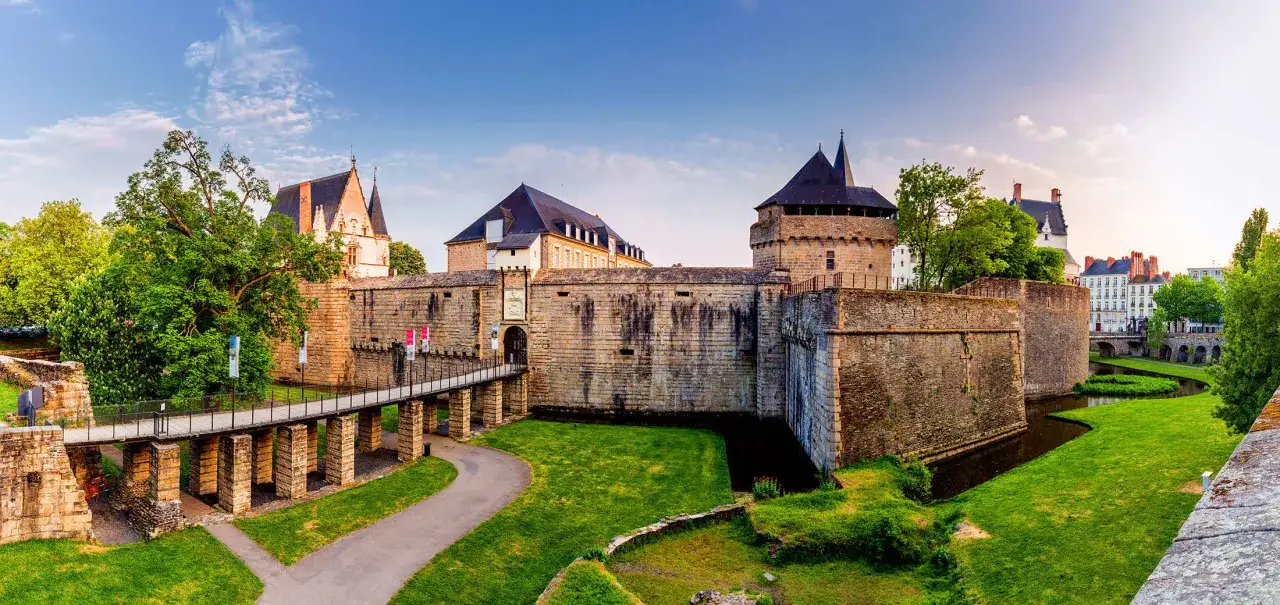Le Ch&acirc;teau de Nantes se dresse fi&egrave;rement, entour&eacute; de douves verdoyantes. Un pont m&egrave;ne &agrave; son entr&eacute;e fortifi&eacute;e, sous un ciel bleu clair.