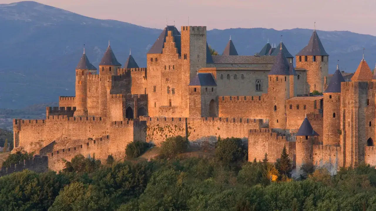 La Cit&eacute; de Carcassonne, un des plus c&eacute;l&egrave;bres ch&acirc;teaux en France, se dresse majestueusement au coucher du soleil, ses remparts dor&eacute;s se d&eacute;tachant sur les collines bleut&eacute;es.