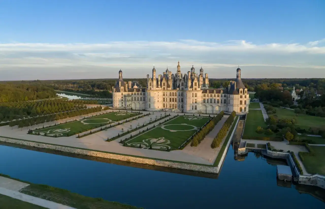 Le majestueux château de Chambord se reflète dans les douves, entouré de jardins à la française et d'une forêt luxuriante sous un ciel bleu clair.