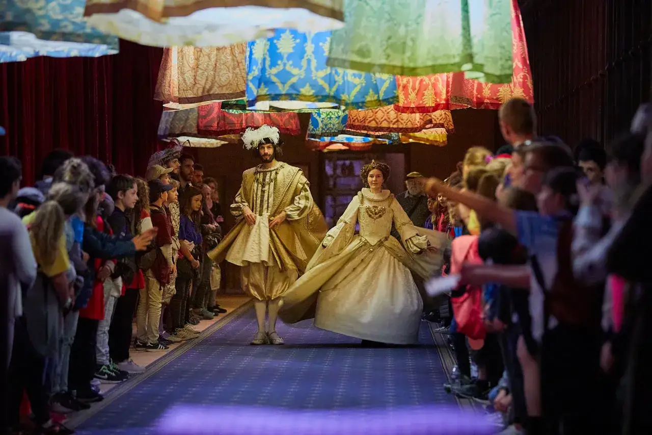 Un couple royal en costumes d'époque défile devant une foule au Puy du Fou, sous des tentures colorées.