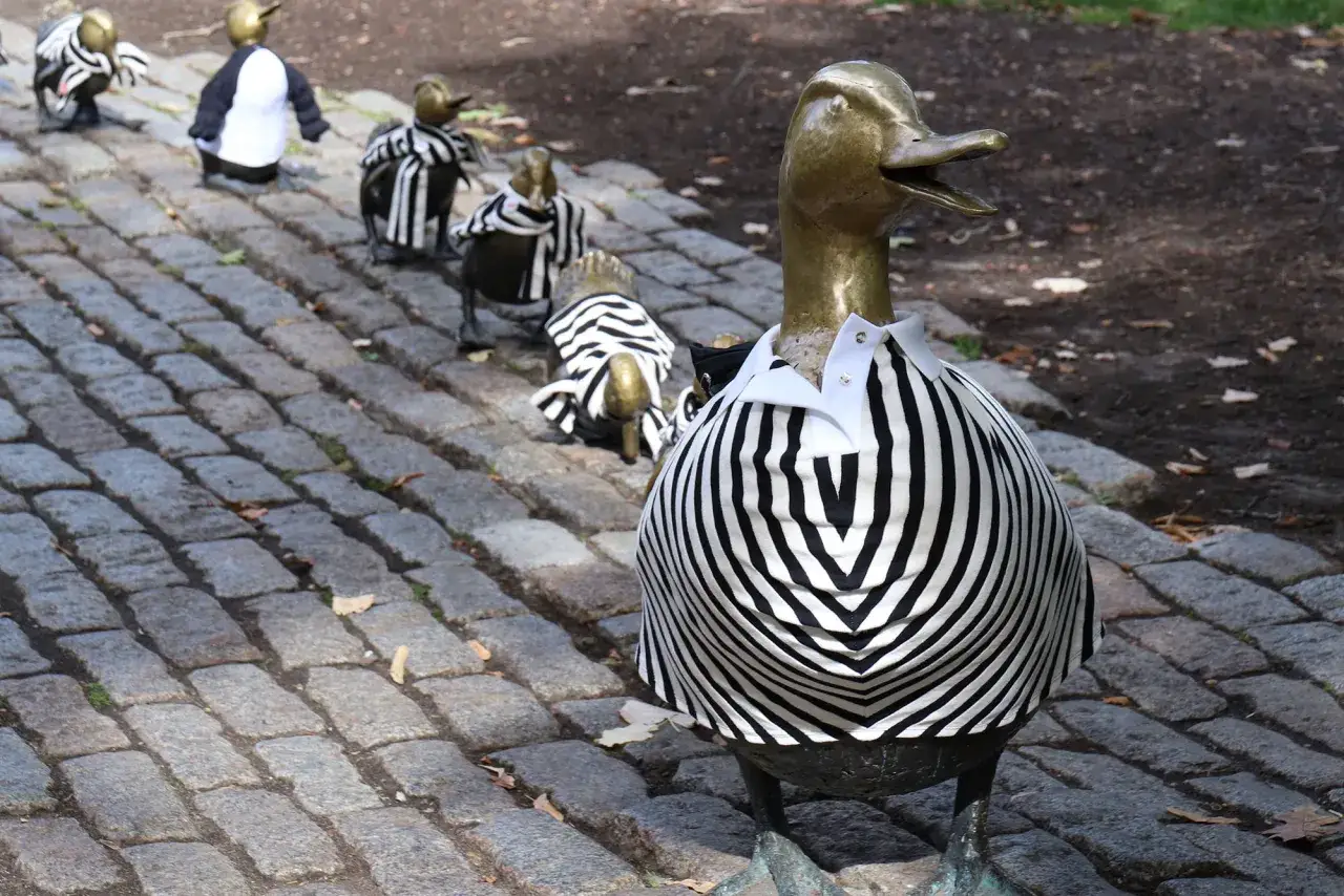 Une joyeuse troupe de canards en bronze, symboles du canard, défilent sur un chemin pavé, vêtus de tenues rayées noir et blanc.