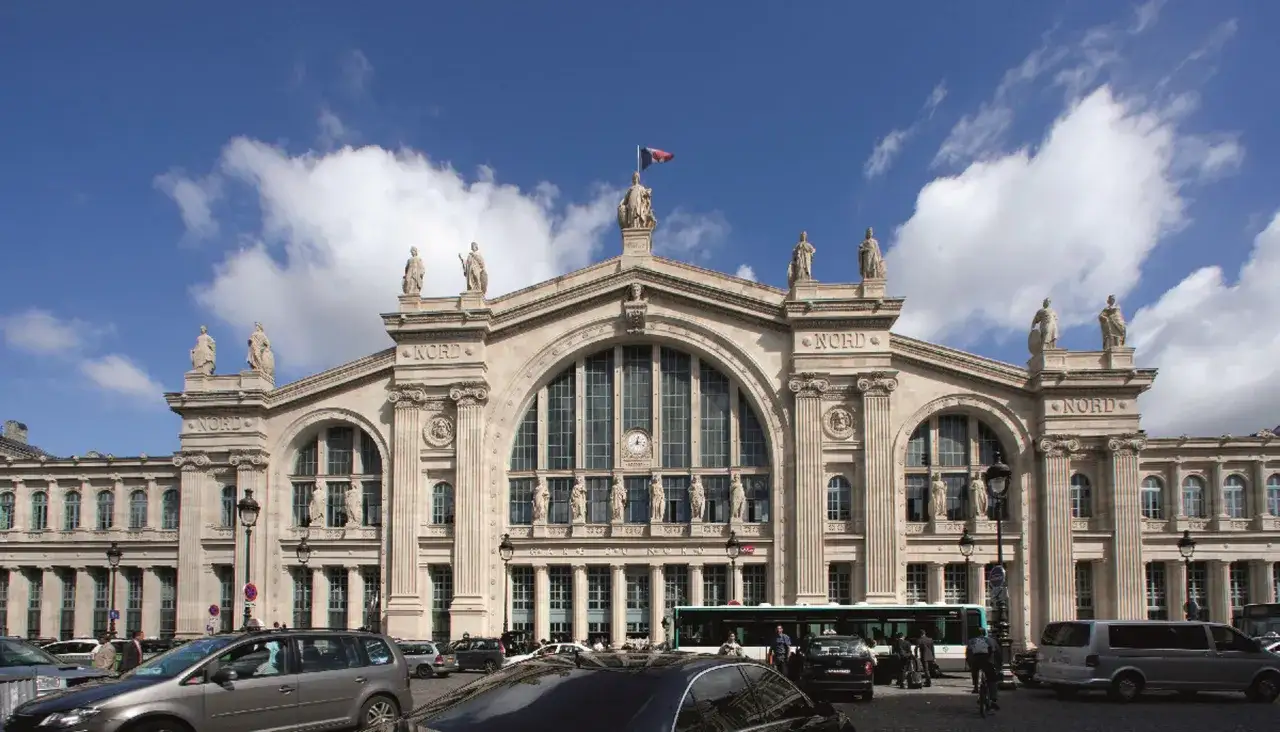 La Gare du Nord à Paris, majestueuse sous un ciel bleu et nuageux, avec ses statues et son architecture imposante. Des voitures et un bus circulent devant.