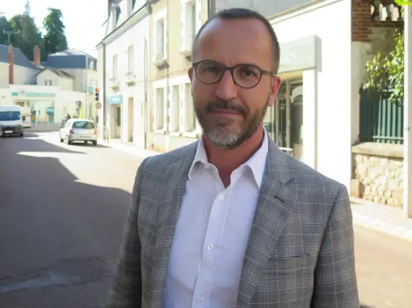 Laurent Raymond, homme politique, porte une veste à carreaux et une chemise blanche, debout dans une rue ensoleillée.